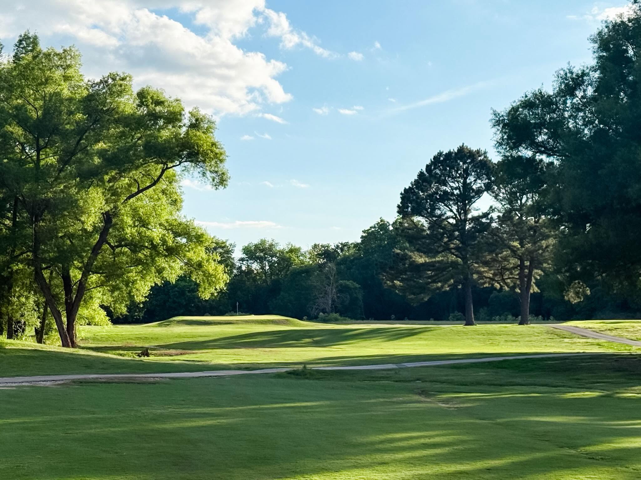 A clean striped fairway with a cart path and big shade trees under blue sky at The Creeks