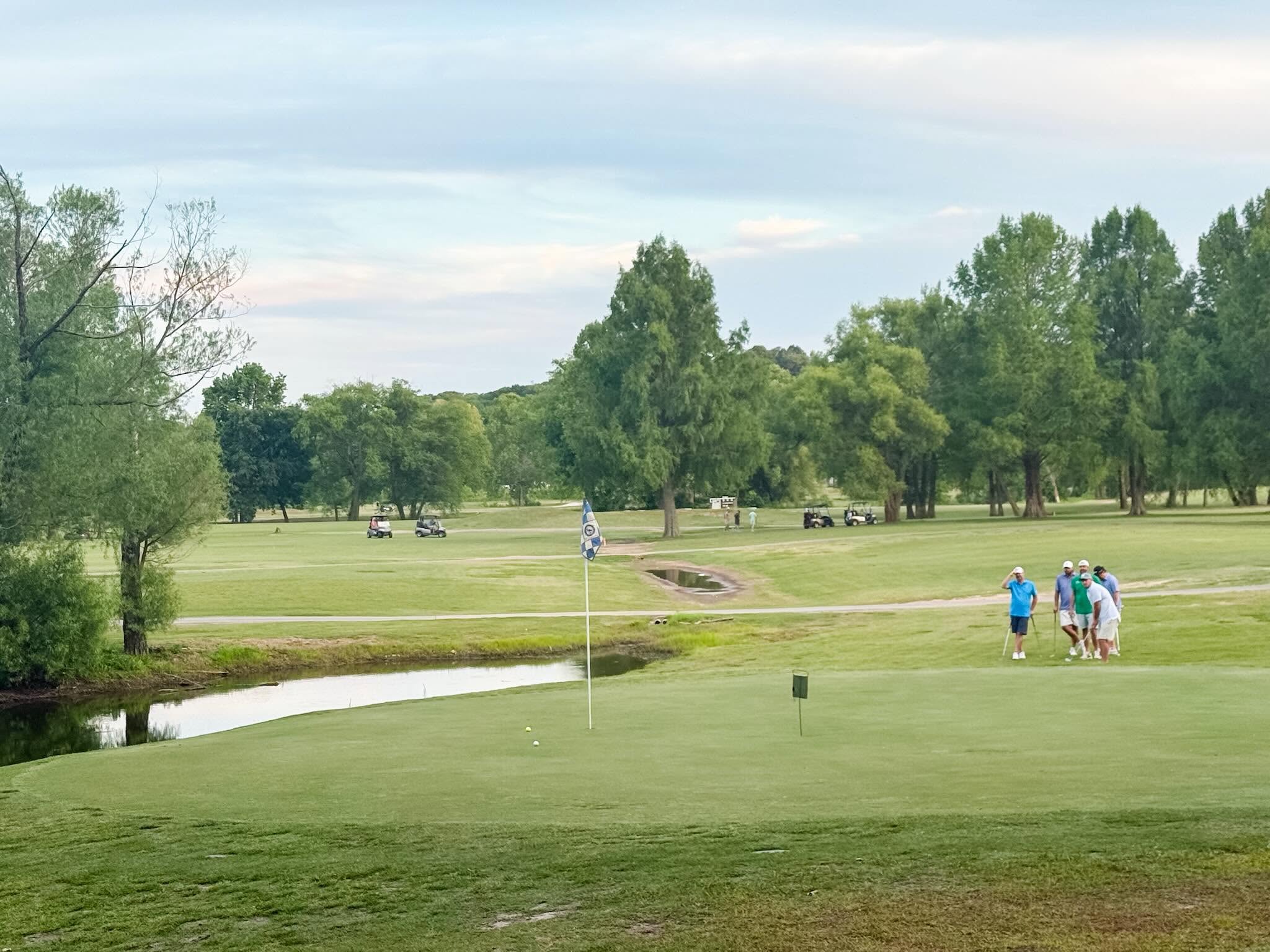 A green guarded by water with players threading the fairway beyond at The Creeks